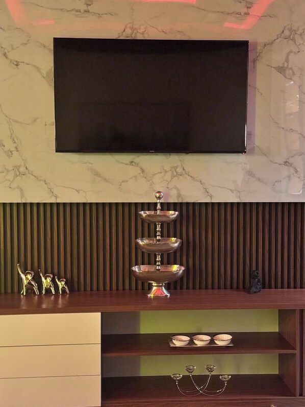 A modern living room with a flat-screen TV mounted on a marble wall, a decorative silver stand, and a wooden cabinet with glassware.