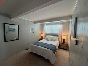 Un dormitorio con cama, mesitas de noche, lámpara, ventana con cortinas y televisor montado en la pared.
