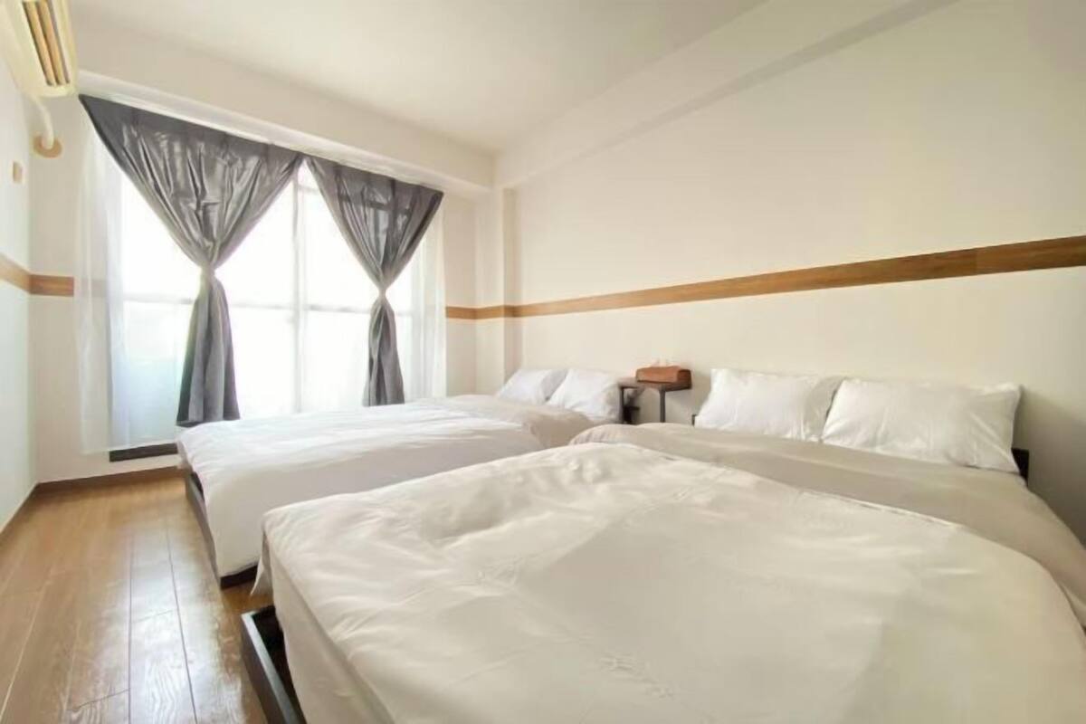 Habitación de hotel con dos camas, piso de madera, ventana con cortinas y mesita de noche con lámpara.