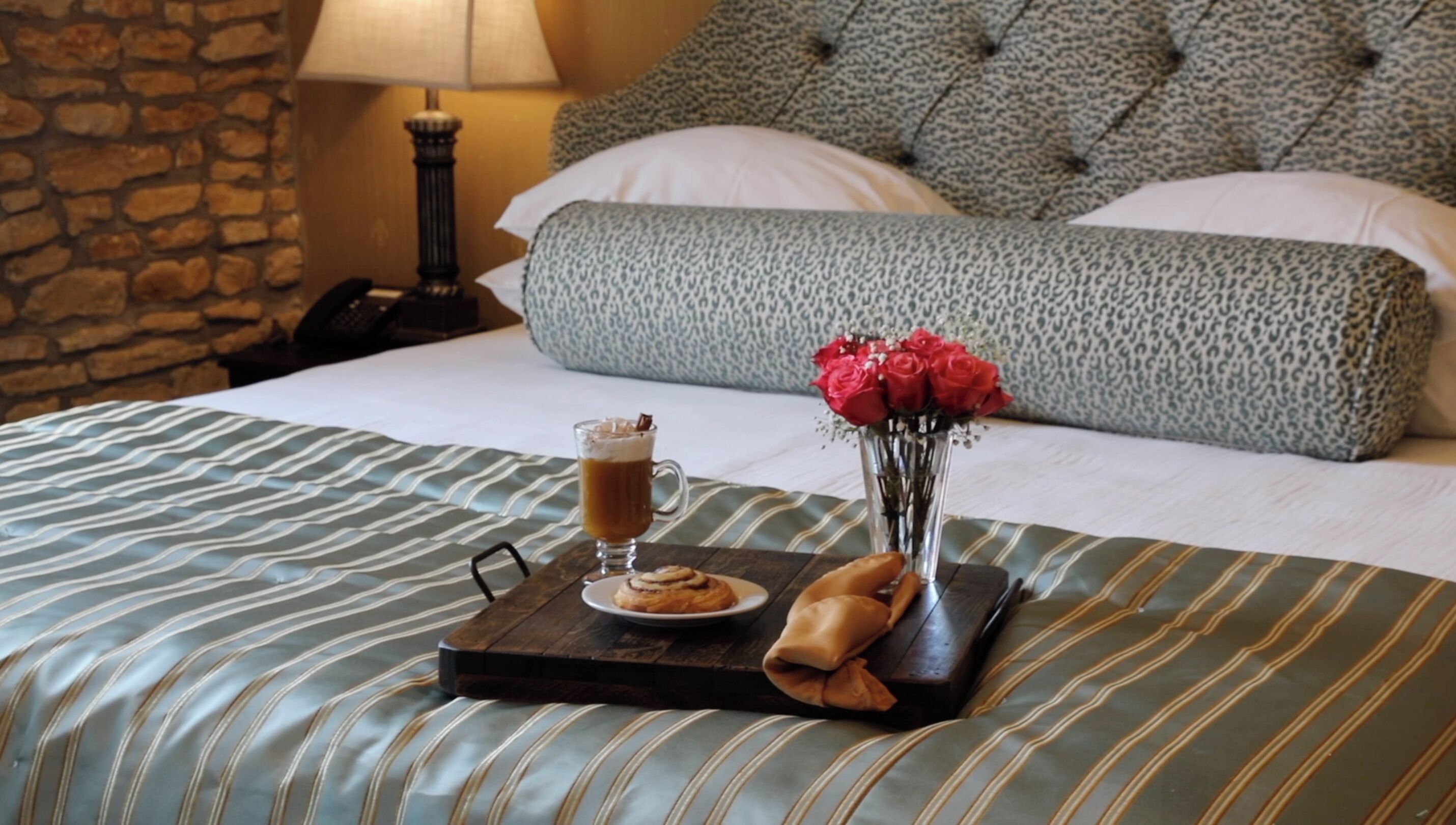 A neatly made bed with striped bedding, a tray with a pastry, a glass of beverage, and a vase of red roses.