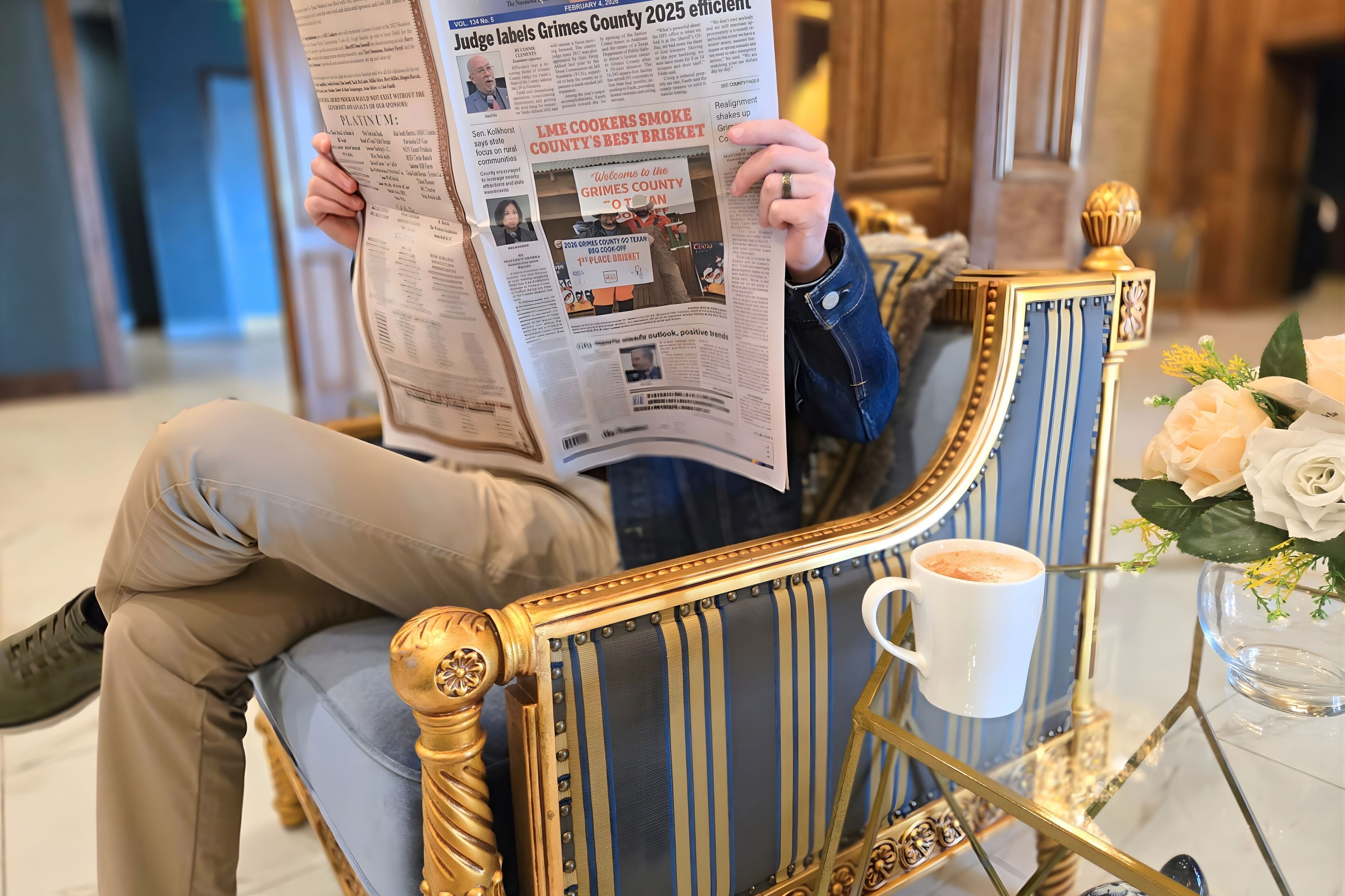 A person reading a newspaper in a room with a decorative chair, a coffee cup, and a vase with flowers.