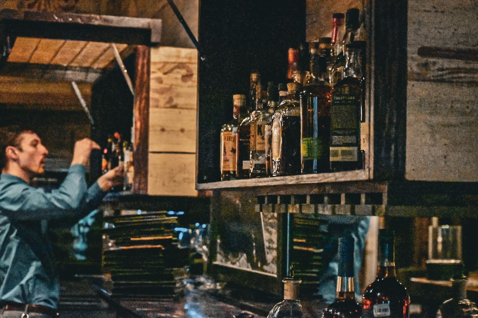 A bar area with shelves stocked with bottles, a person holding a bottle, and a wooden structure in the background.