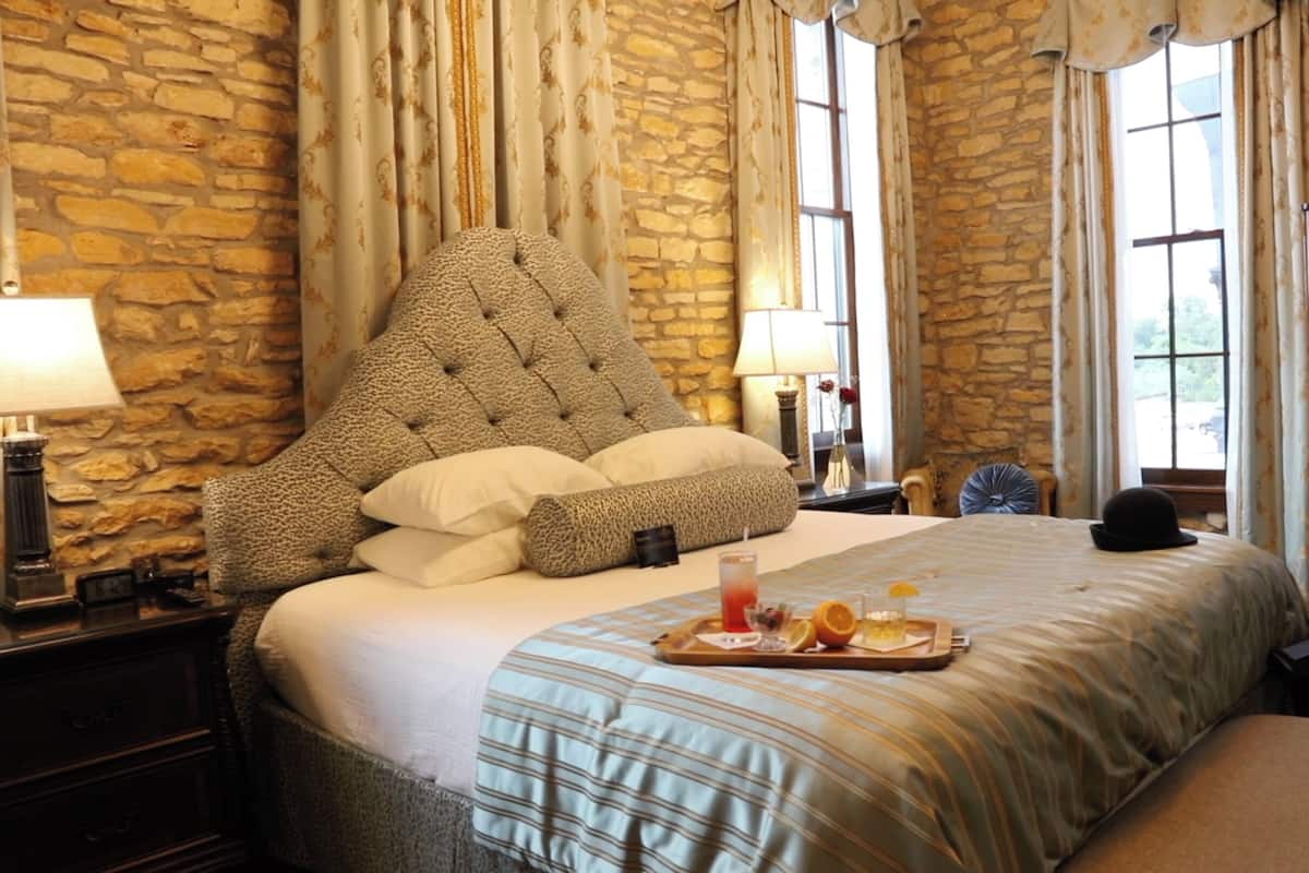 A hotel room with a stone wall, a tufted headboard, a bed with a striped bedspread, and a tray with fruit and a drink on the bedside table.