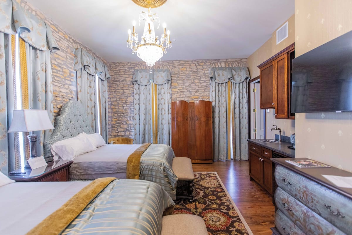A hotel room with a stone wall, a chandelier, a bed with two pillows, a bench, a wooden wardrobe, a dresser, and a television.