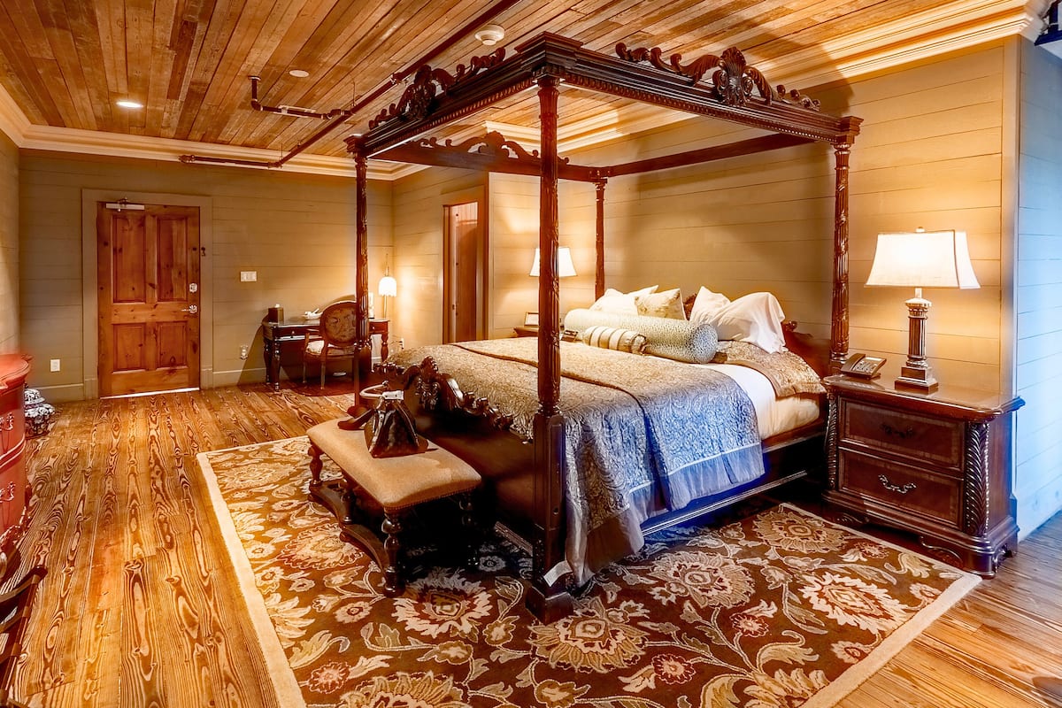 A four-poster bed with a wooden canopy, a bench, and a nightstand with a lamp.