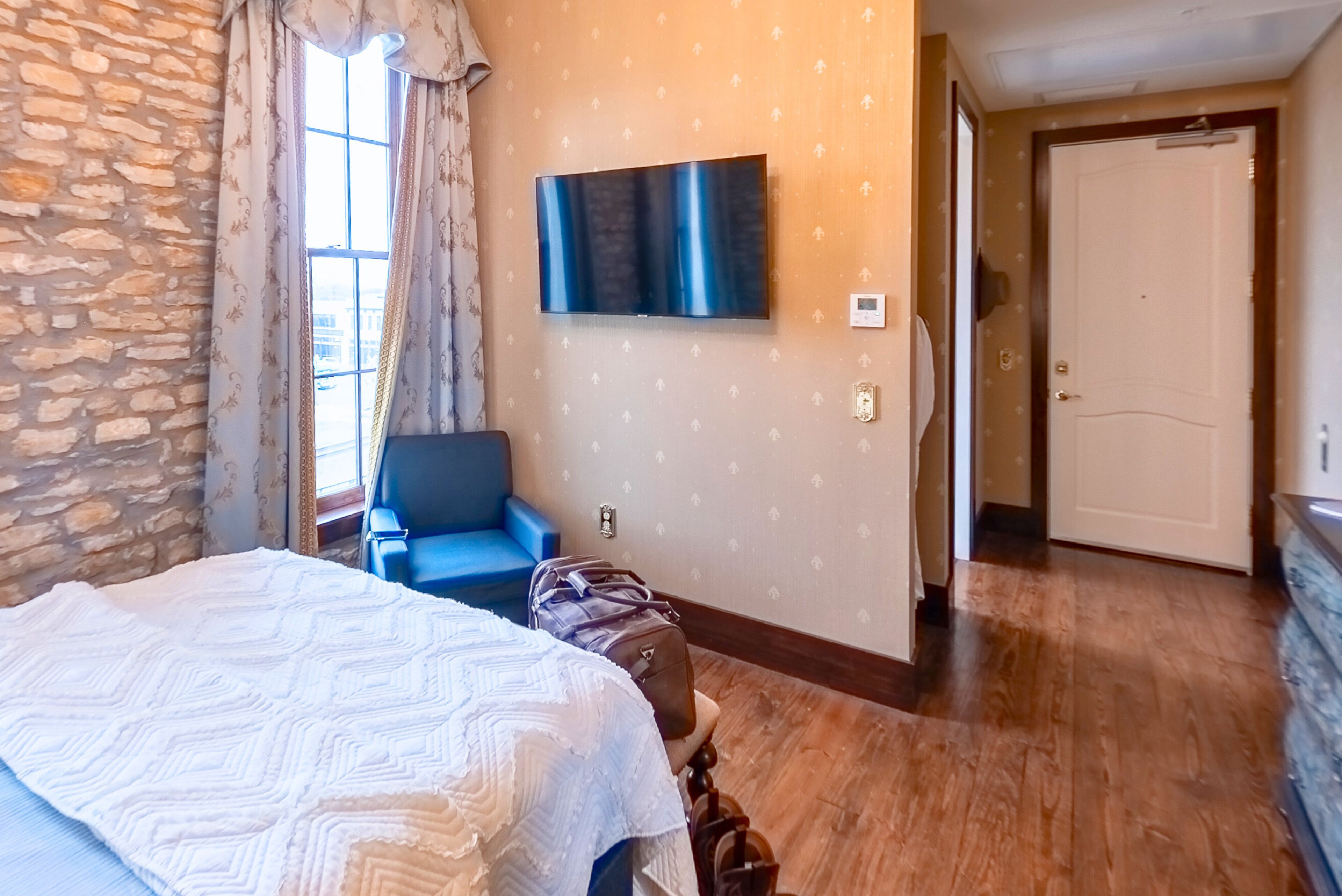 A hotel room with a bed, a TV mounted on the wall, a blue chair, and a wooden floor.