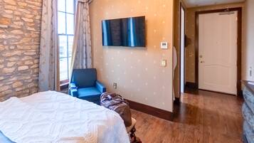 A hotel room with a bed, a TV mounted on the wall, a blue chair, and a wooden floor.
