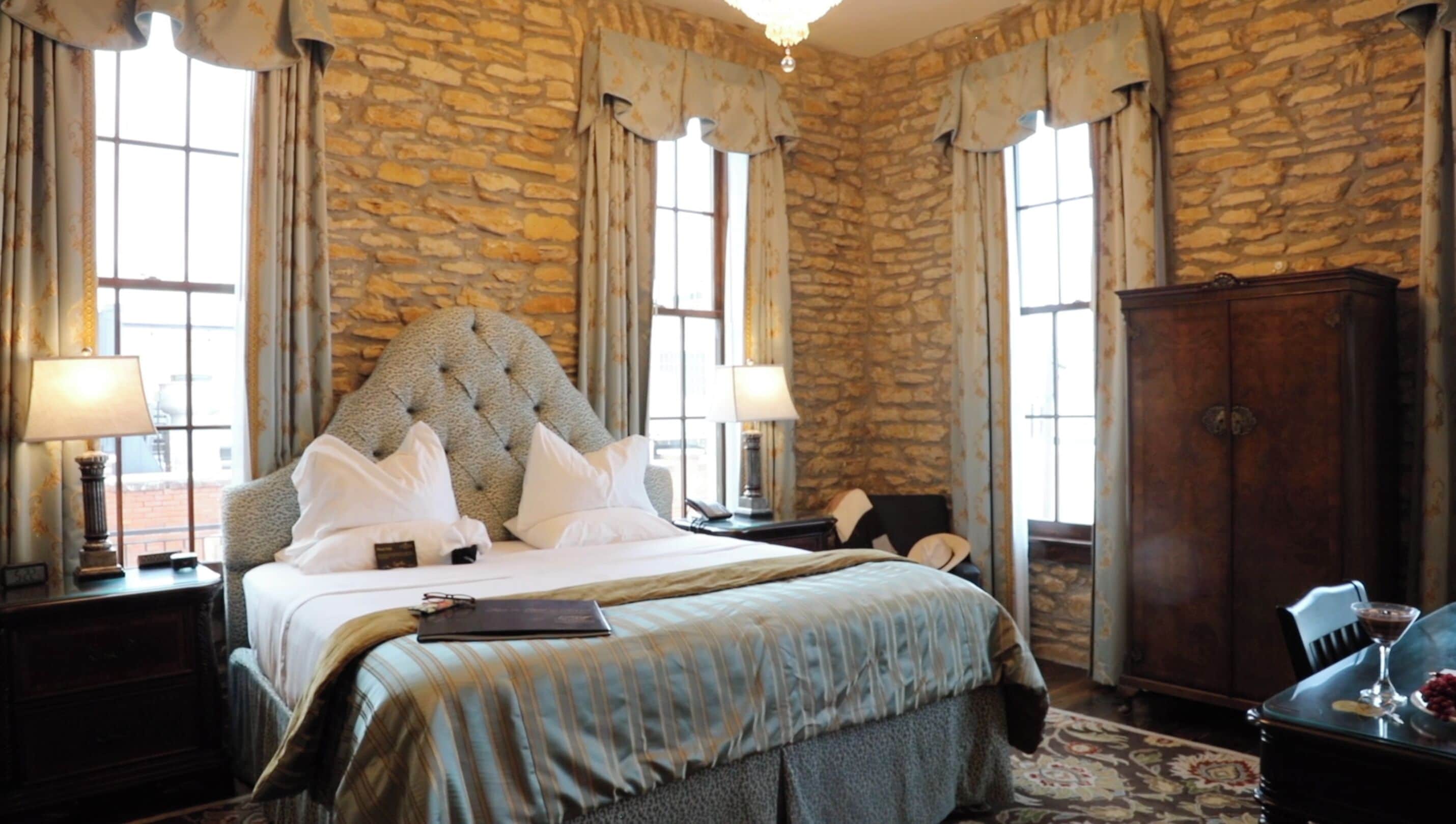 A bedroom with a large bed, stone walls, and two windows with curtains.