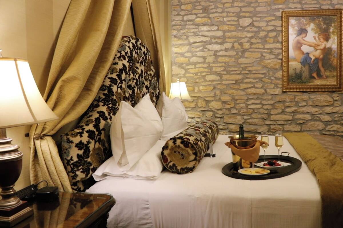 A hotel room with a stone wall, a bed with a canopy, a painting, and a tray with food and drinks.