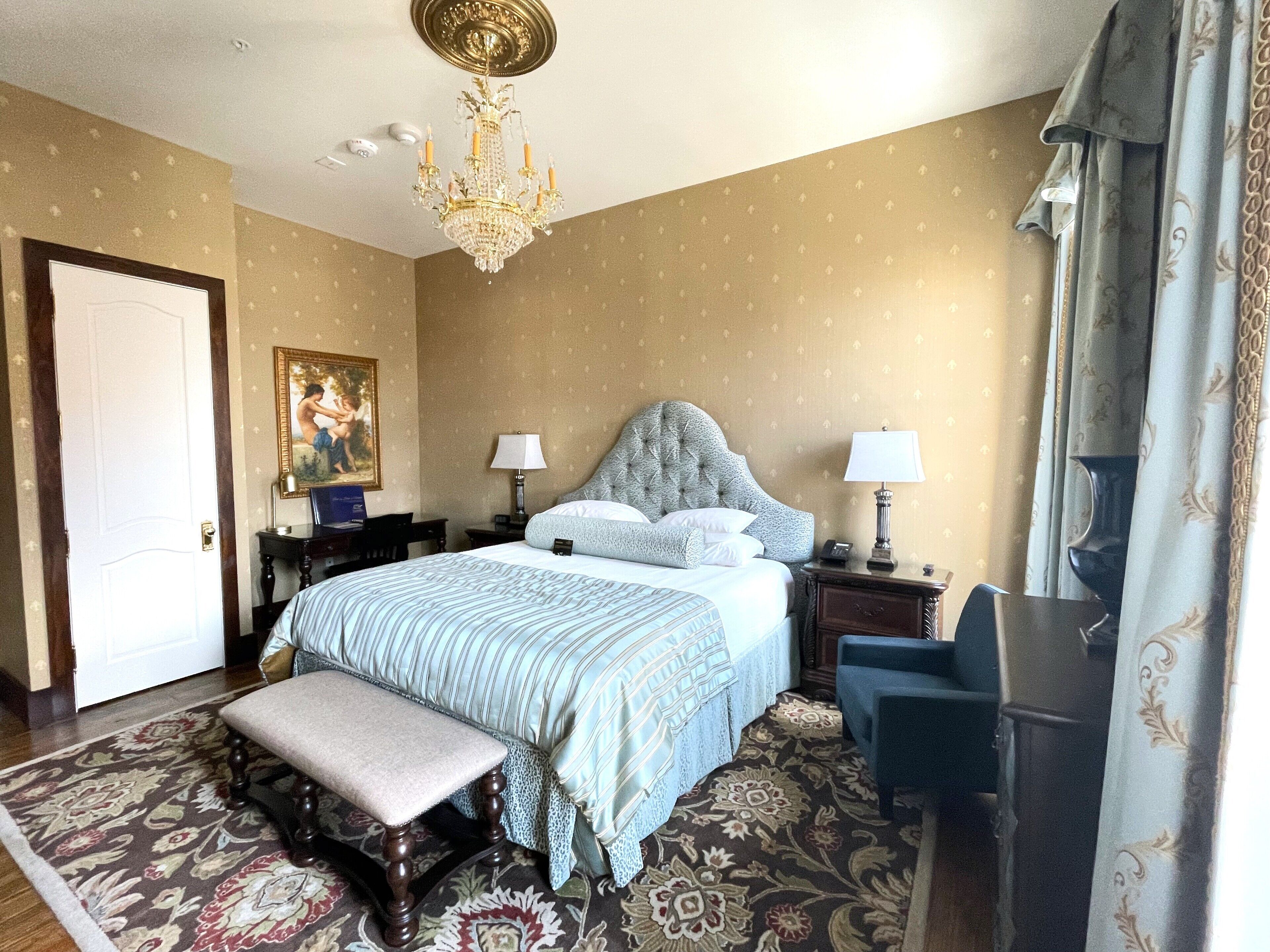 A hotel room with a large bed, two bedside tables, a desk, a chair, a bench, a chandelier, and a door.
