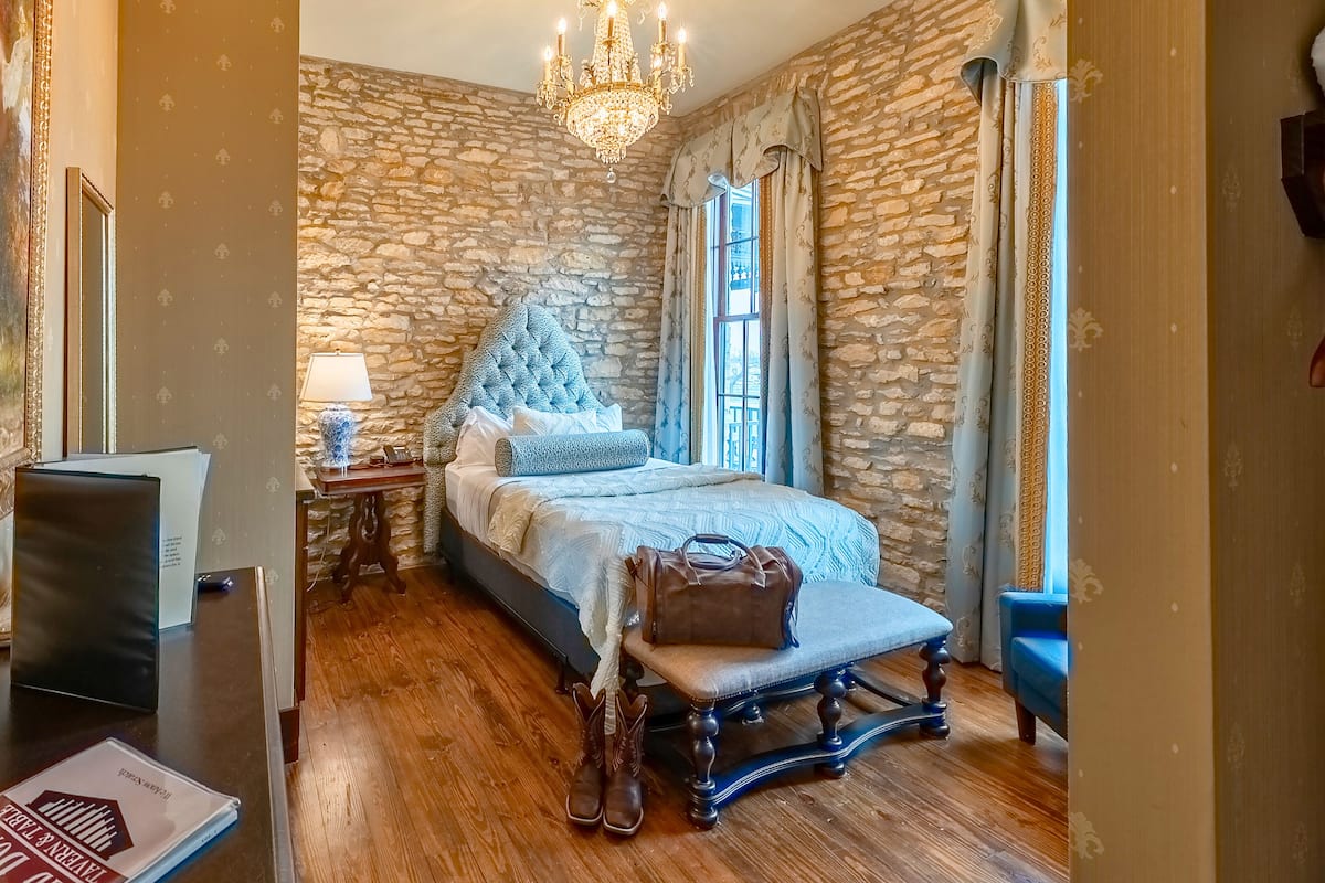 A bedroom with a stone wall, a chandelier, a bed with a blue headboard, a bench with a handbag, and a desk with a lamp.