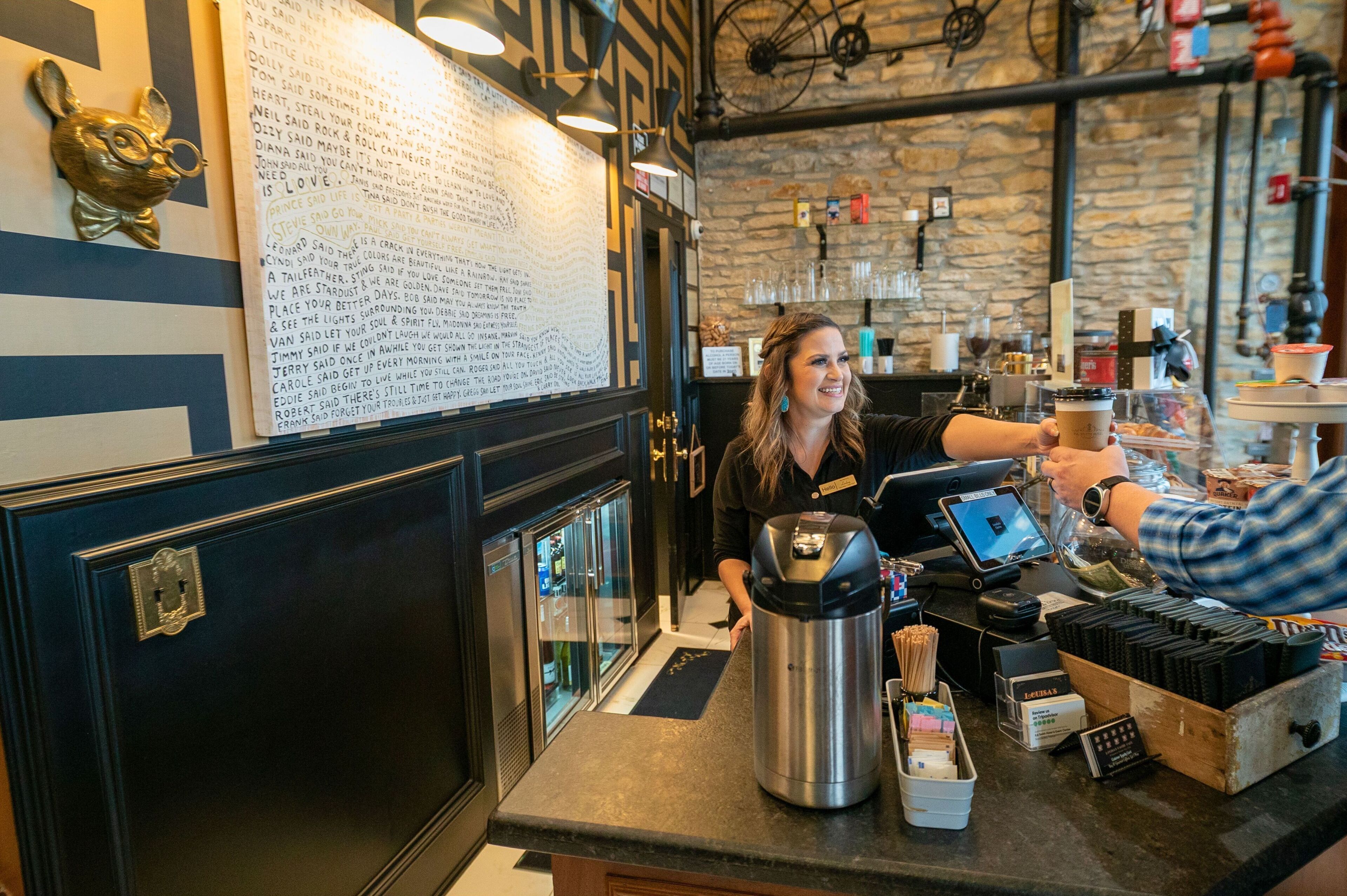 Eine Barista bereitet ein Getränk an der Theke zu, mit einer Kaffeemaschine, einer Speisekarte und einer Steinwand im Hintergrund.
