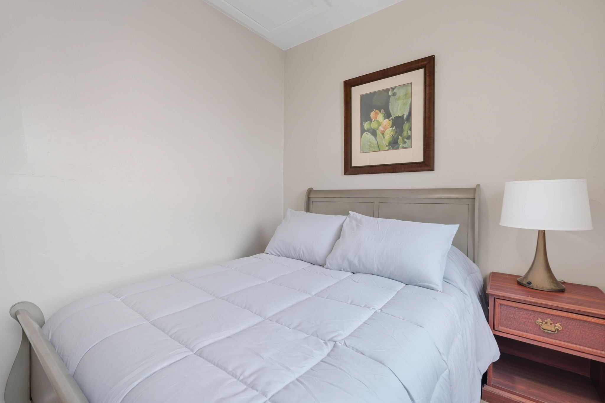 A neatly made bed with a light-colored duvet, a wooden nightstand with a lamp, and a framed picture on the wall.