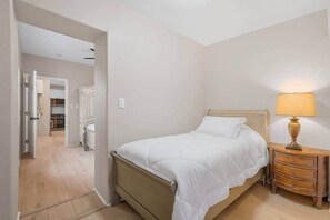 Ein Schlafzimmer mit Bett, Nachttisch und Lampe. Eine Tür führt zu einem Badezimmer und einem Flur.