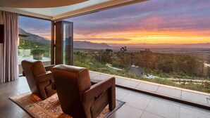 Ein modernes Zimmer mit einer großen Glasdoor, die auf einen Balkon mit herrlichem Bergblick und Sonnenuntergang führt.