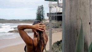 A person in a bikini by a wooden post with a showerhead, overlooking a beach and ocean.