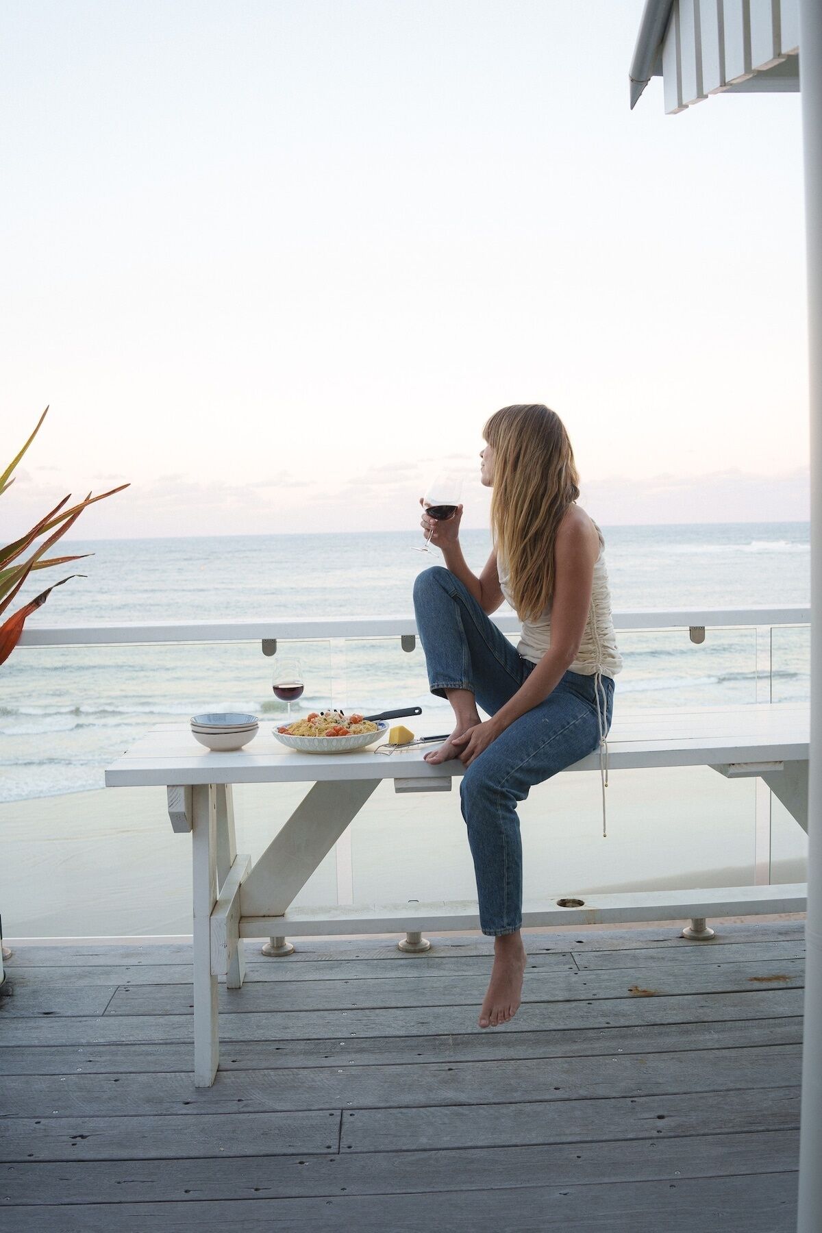 A person sitting at a white table with a bowl of food and a glass of red wine, overlooking the ocean.