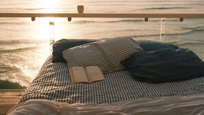 A bed with a book and a cup, overlooking a beach with the ocean and a sunset.