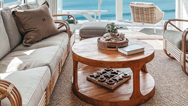 A cozy living room with a large window overlooking the ocean, a round wooden coffee table, wicker furniture, and a view of the sea.