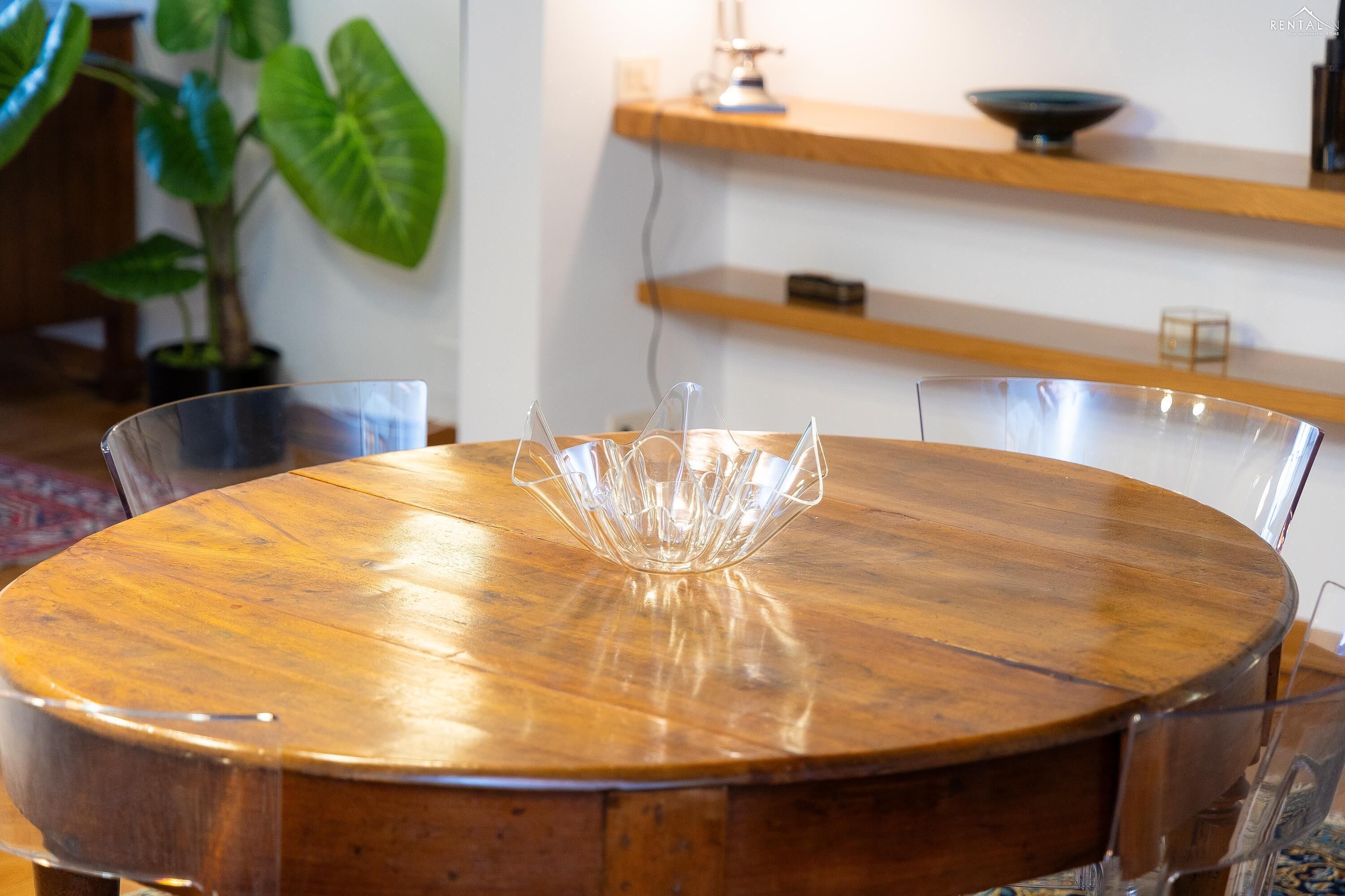 A round wooden dining table with a clear glass bowl in the center, surrounded by metal chairs.