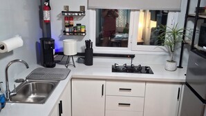 A compact kitchen with a sink, stove, and cabinets. There is a window with a view of the outside, a shelf with kitchen items, and a plant on the counter.