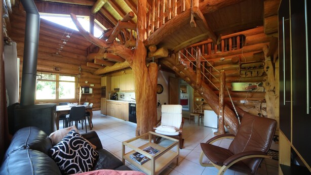 Interior de una cabaña de troncos con comedor, cocina y una escalera que sube a un nivel superior.