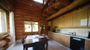 Interior de una cabaña de madera con una mesa de comedor, sillas, una cocina con armarios de madera y una ventana.
