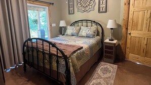 Un dormitorio con cama, ventilador de techo, mesita de noche, lámpara, puerta y ventana con cortinas.