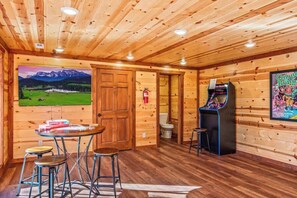 A wooden cabin interior with a dining area, a game machine, and a mountain view painting. - 6-Bedroom Cabin with Indoor Pool, Movie Theater & Hot Tub Near Dollywood (Sevierville)