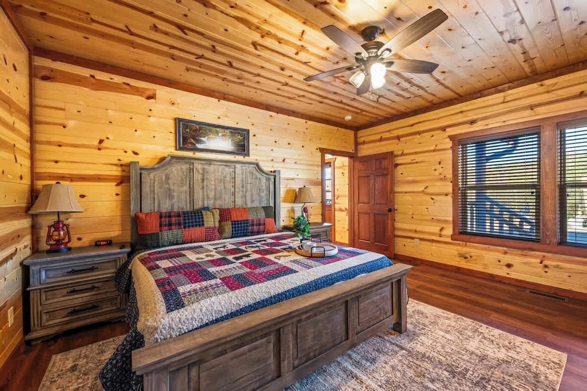 Une chambre à coucher dans une cabane en bois, avec un lit, des tables de chevet, un ventilateur de plafond et une fenêtre avec des stores.