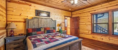 Un dormitorio de cabaña de madera con cama, mesitas de noche, ventilador de techo y ventana con persianas.