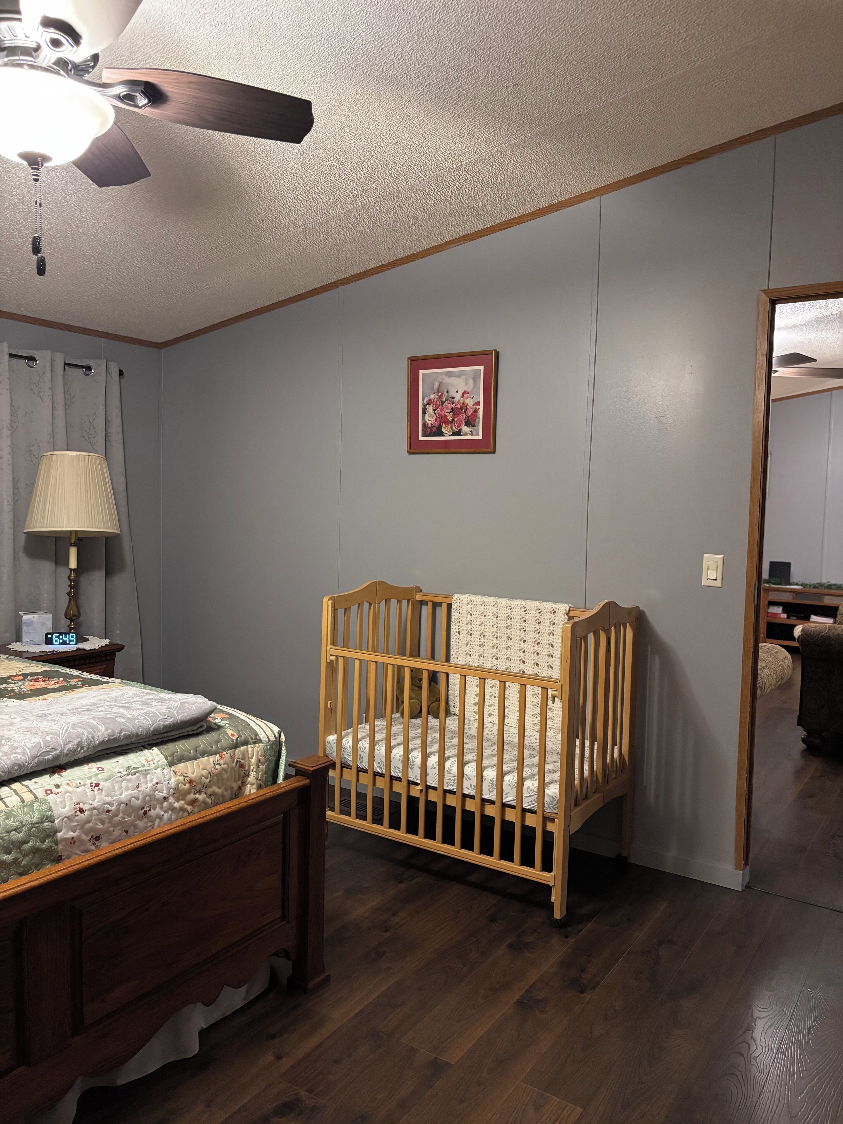 A bedroom with a wooden crib, a bed with a floral bedspread, a nightstand with a lamp, and a ceiling fan.
