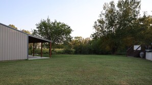 A grassy backyard with a covered patio area, a metal building, and trees in the background. - Beautiful home with pool in Wayne City (Wayne City)