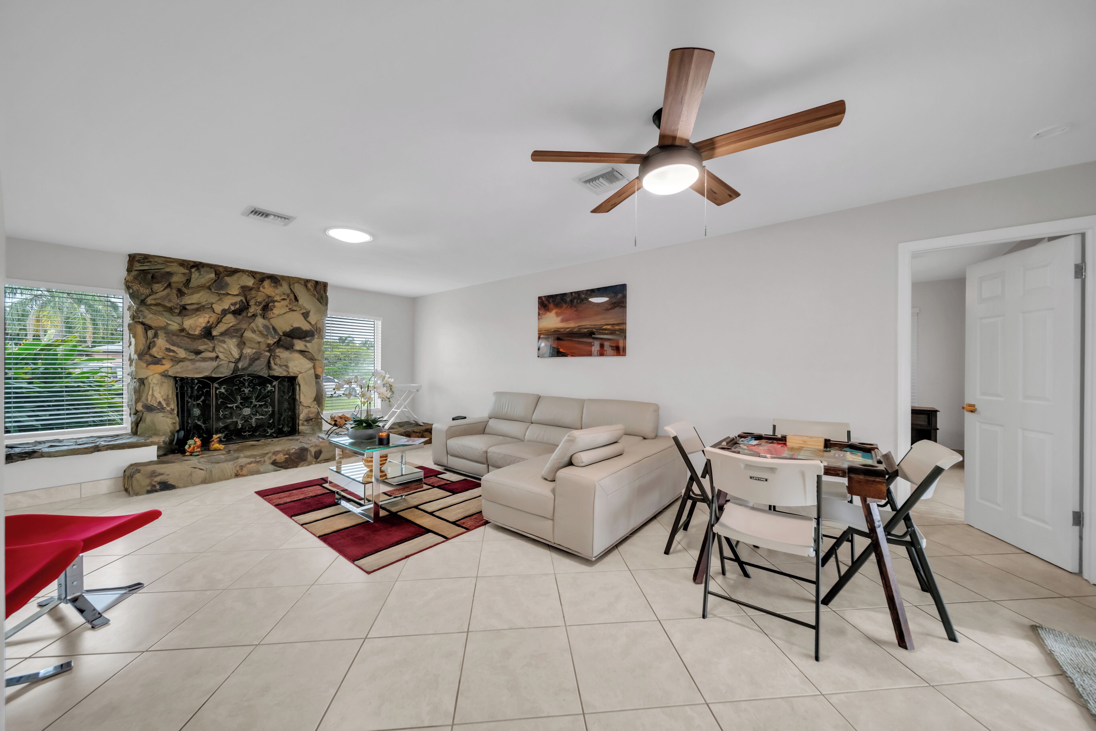 A living room with a stone fireplace, a beige sofa, a dining table with chairs, a ceiling fan, and a door.