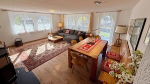 Un salon spacieux comprenant un canapé, une table à manger et une table basse. De grandes fenêtres sont agrémentées de rideaux, et on y trouve également un ventilateur de plafond ainsi qu’une lampe sur une table d’appoint.