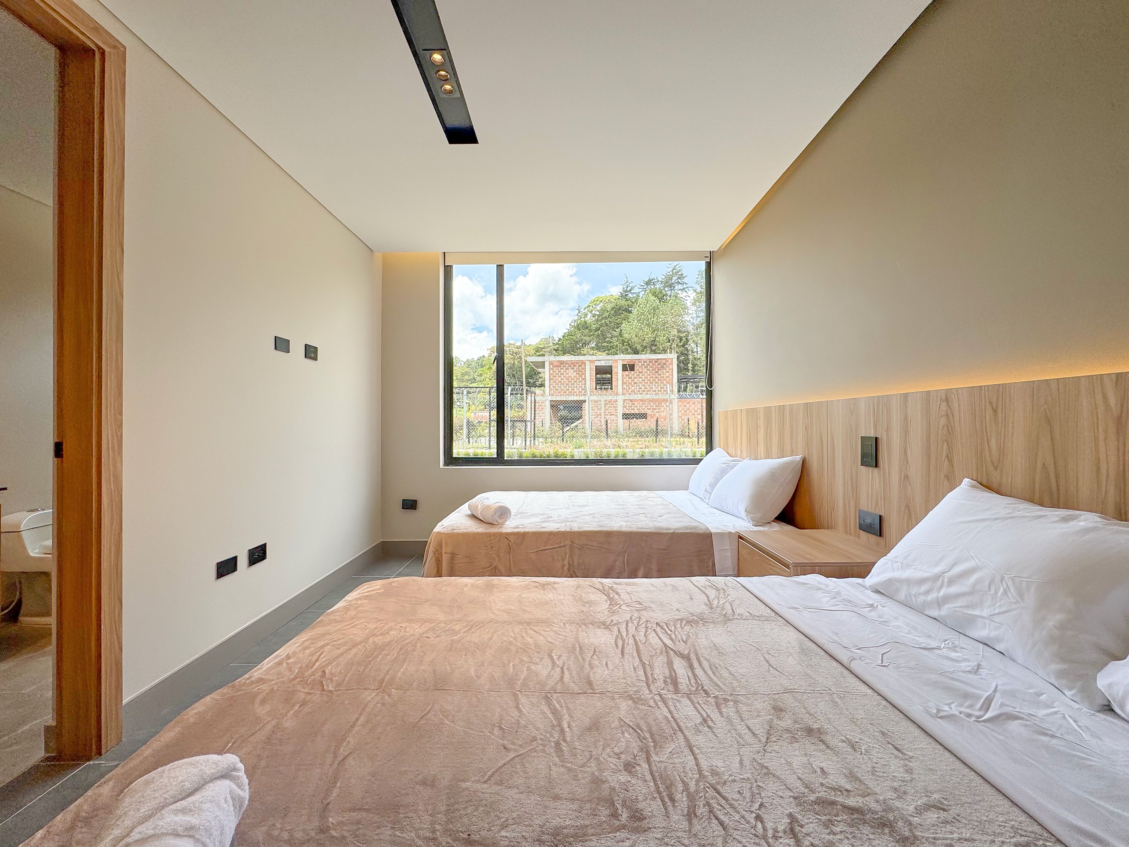 Una habitación de hotel moderna con dos camas, un ventanal grande que ofrece vistas a un edificio de ladrillo y vegetación, y un cabecero de madera.