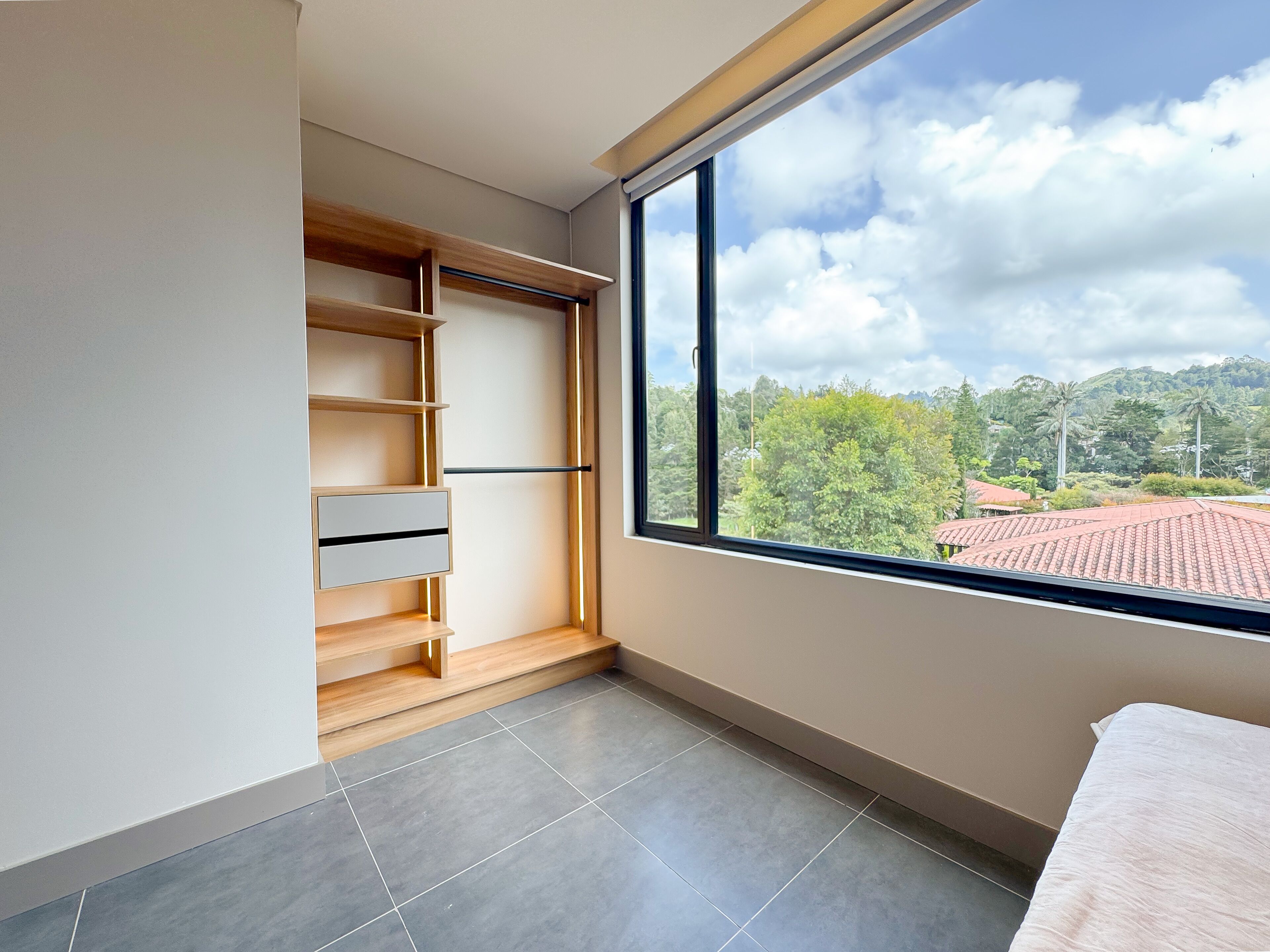 A modern bedroom with a large window, a built-in wooden wardrobe, and a tiled floor.