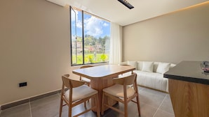 Un comedor moderno con una mesa y sillas de madera, un sofá, y un amplio ventanal con vistas a zonas verdes y edificios.