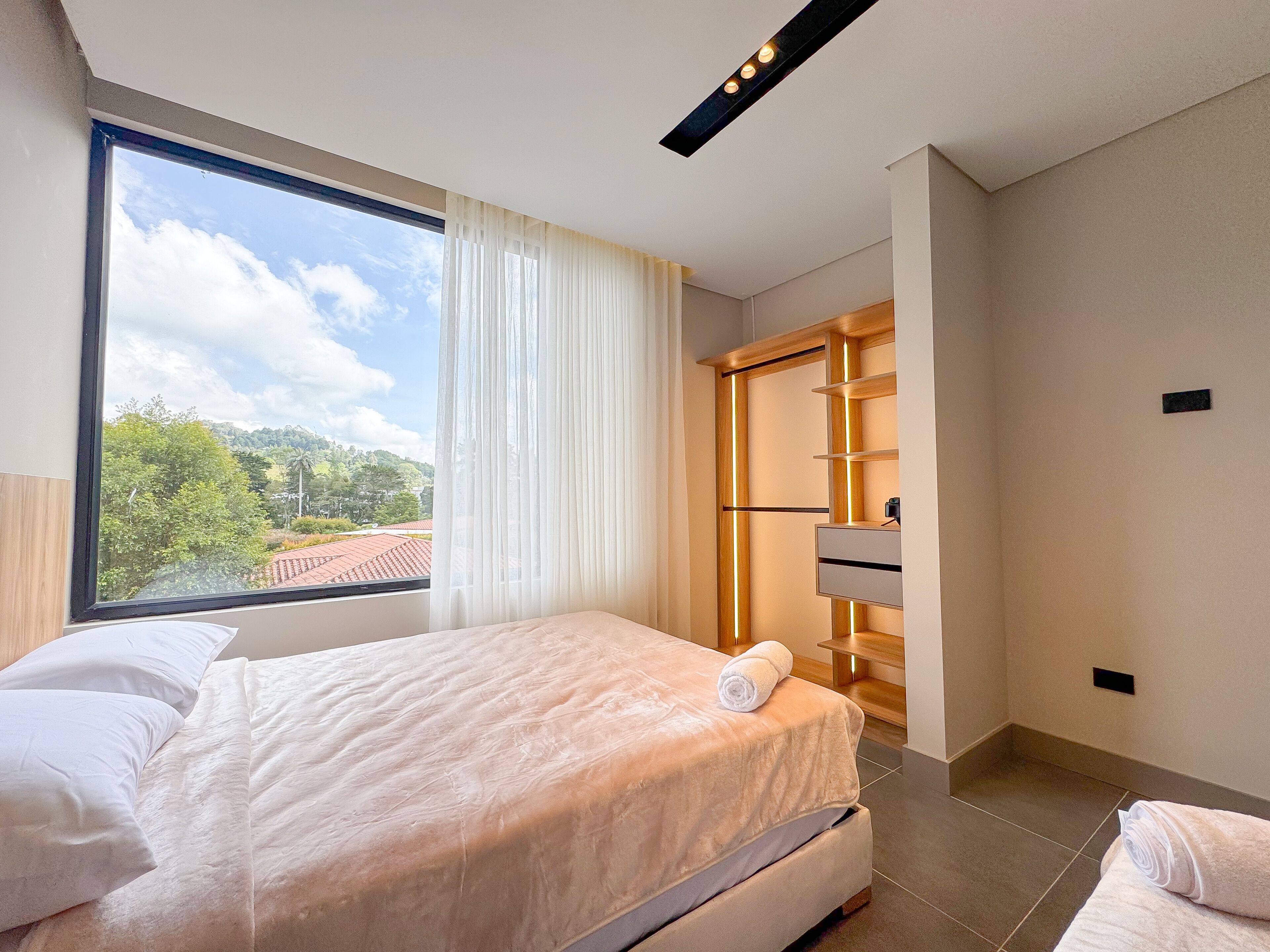 A modern bedroom with a large window, a neatly made bed, a built-in wardrobe, and a view of a green landscape.