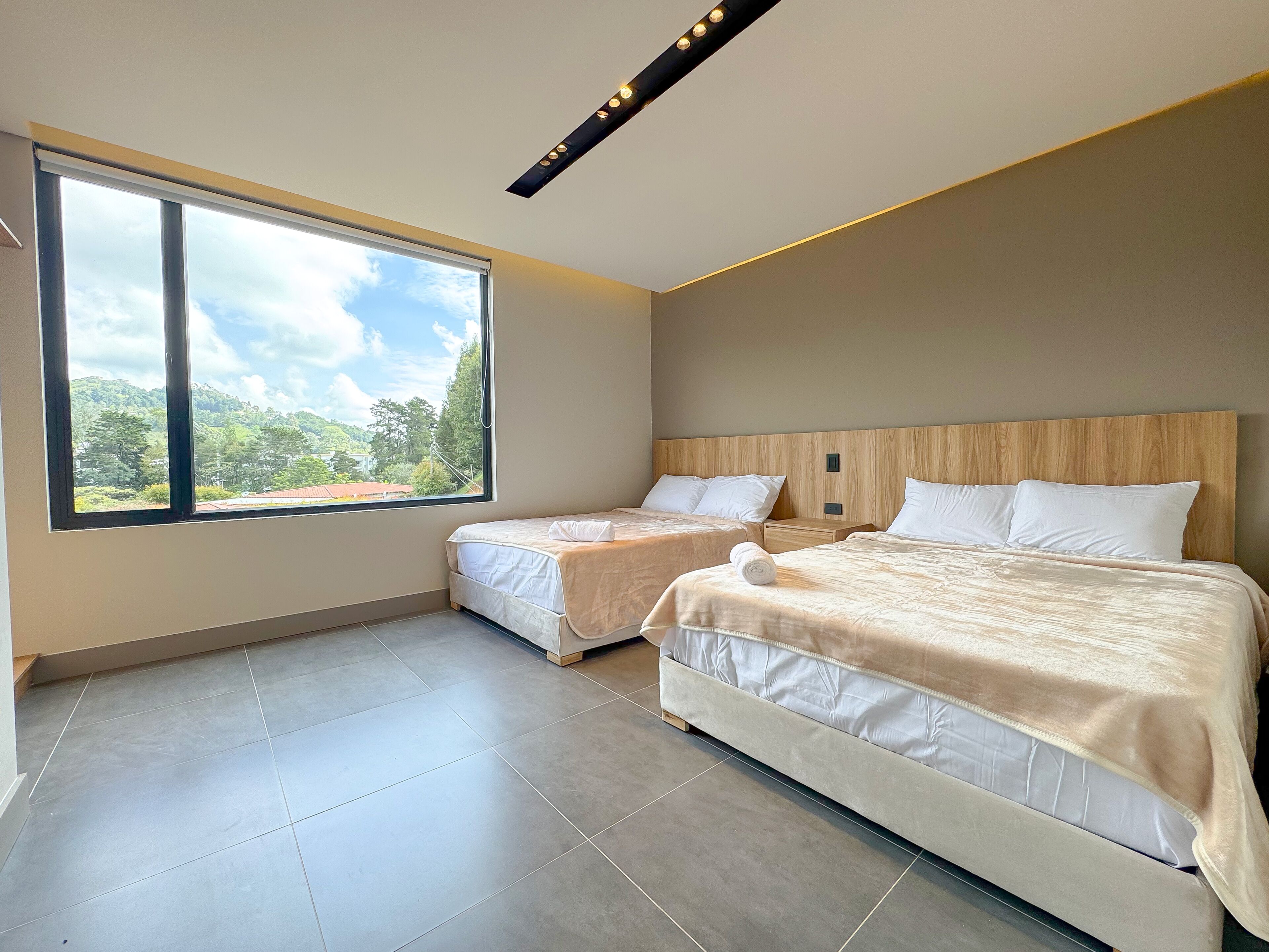 A modern hotel room with two beds, a large window, and a view of greenery.