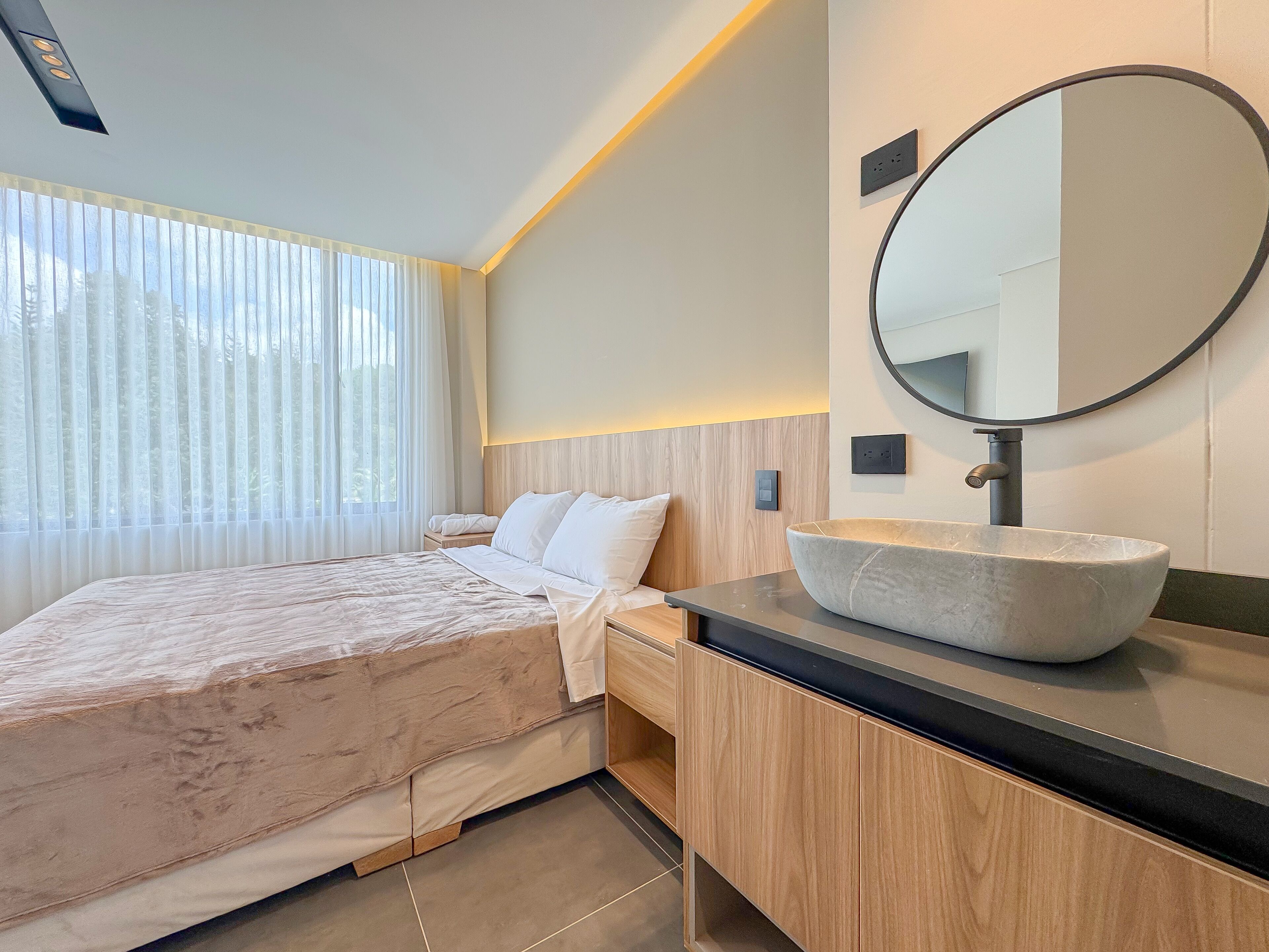A modern bedroom with a large bed, a wooden headboard, a bedside table, a round mirror, and a large vessel sink.