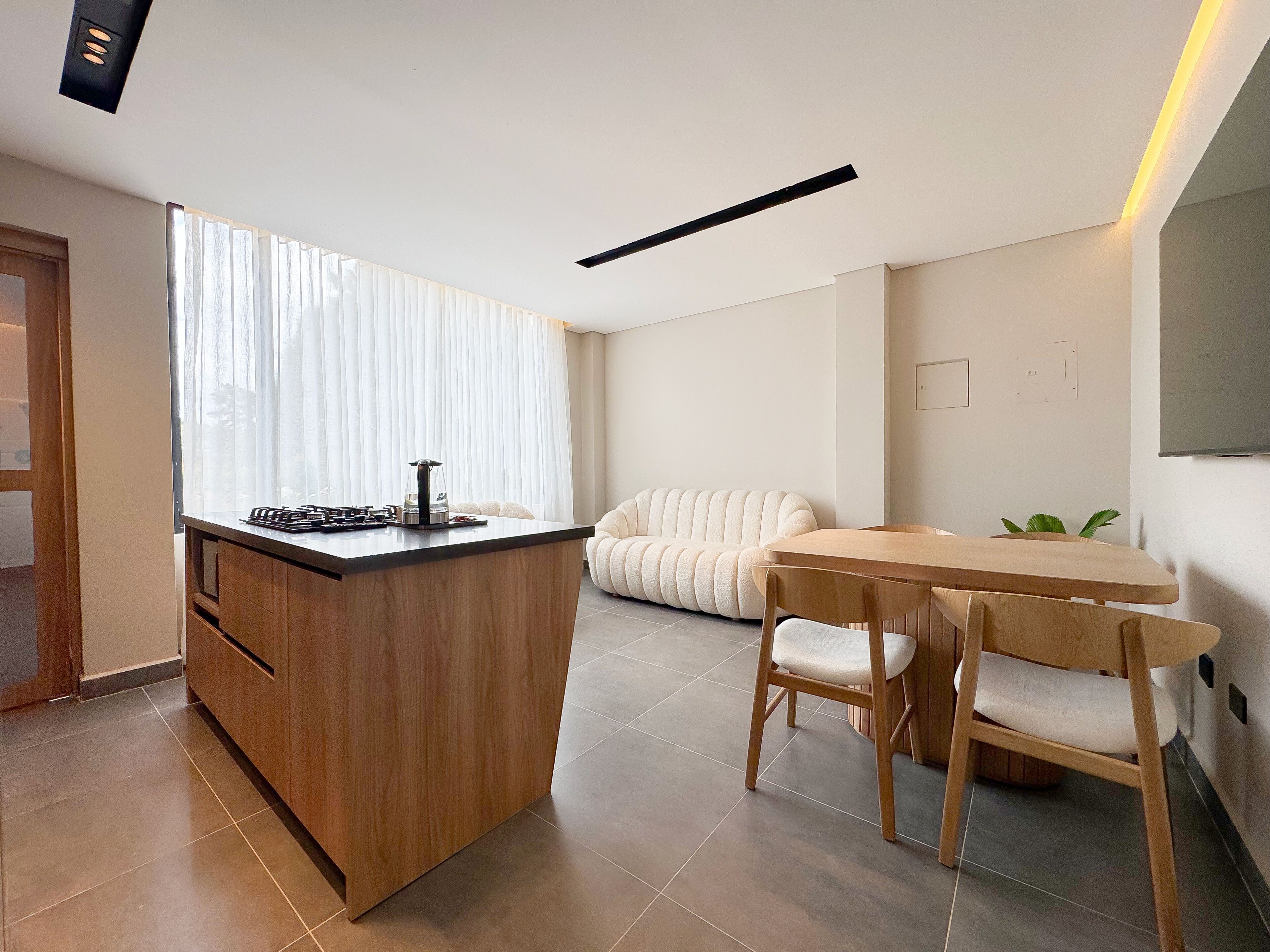 Una cocina moderna con una isla de madera, un comedor con mesa y sillas de madera, y un amplio ventanal con cortinas traslúcidas.