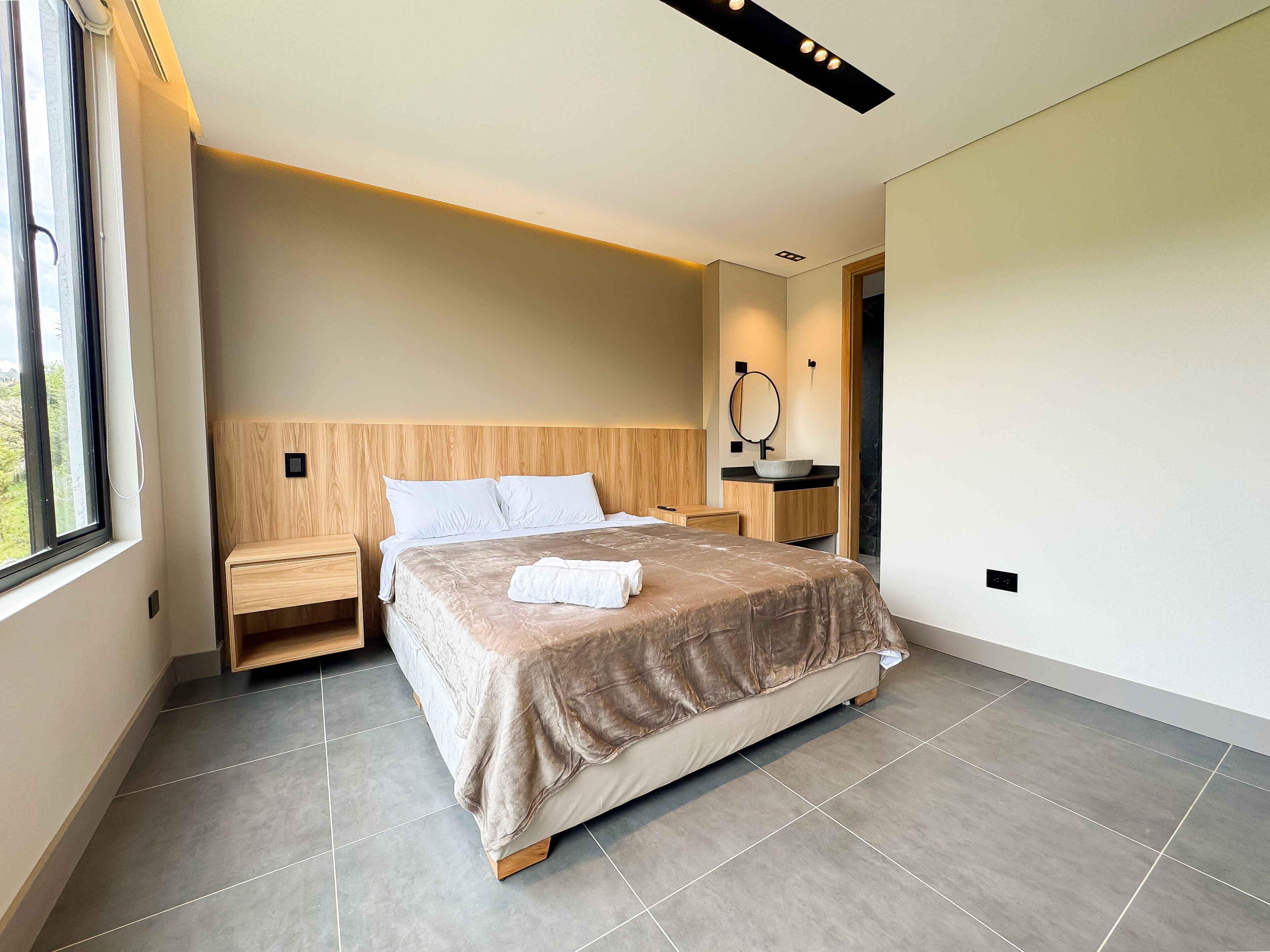 A modern bedroom with a large bed, wooden headboard, bedside tables, a built-in shelf, and a large window.
