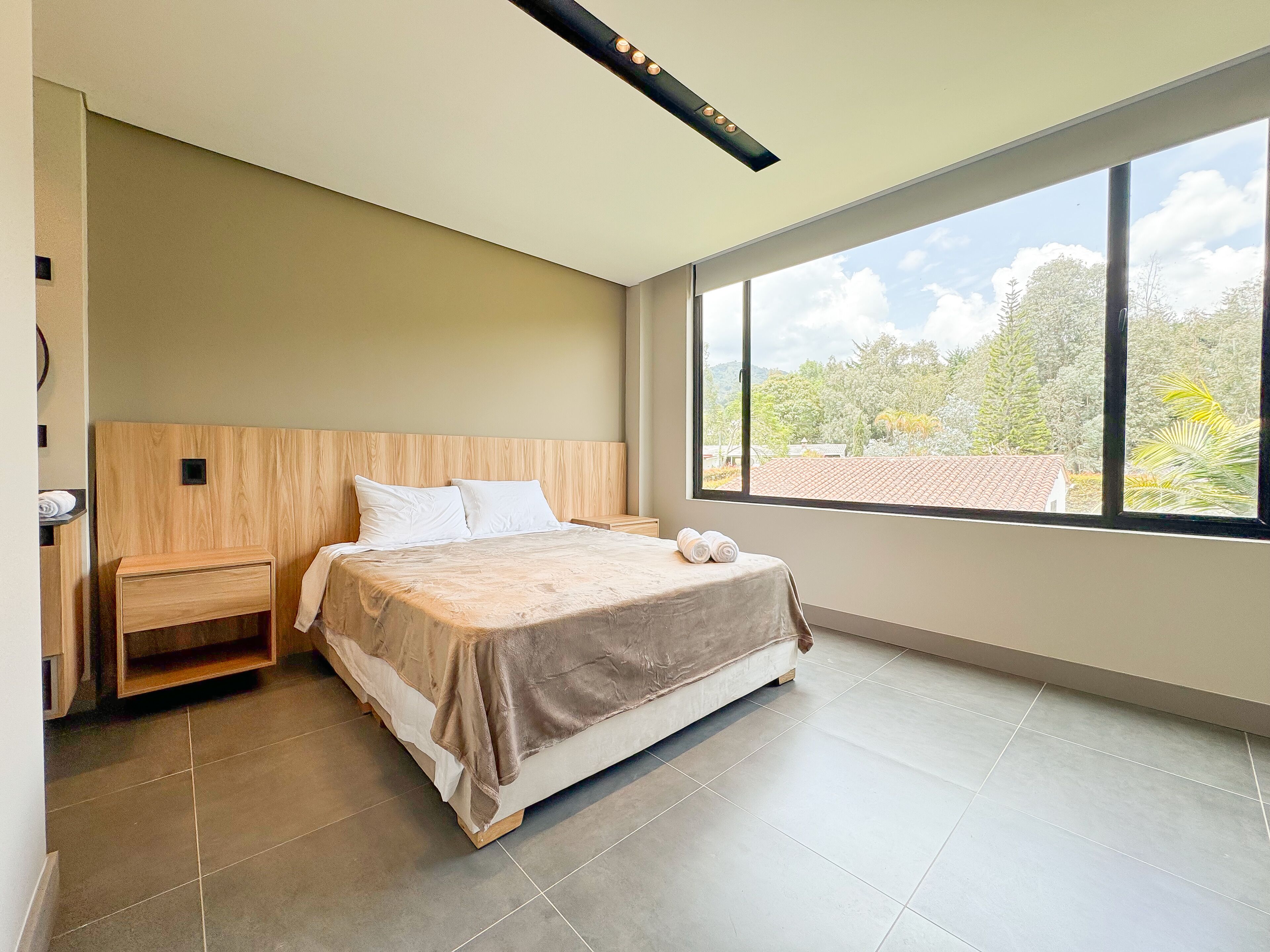 Una camera da letto moderna con una grande finestra, una testiera in legno, un letto con copripiumino color beige e un comodino con lampada.