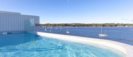 A swimming pool with clear blue water, surrounded by a white pool deck, and a view of the sea with sailboats in the distance.