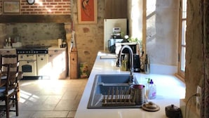 A kitchen with a long counter, a sink, and a stove. There is a clock on the wall and a window providing natural light. - Holiday Home 'L'ancien Collège' with Private Pool, Wi-Fi and Air Conditioning (Hérisson)