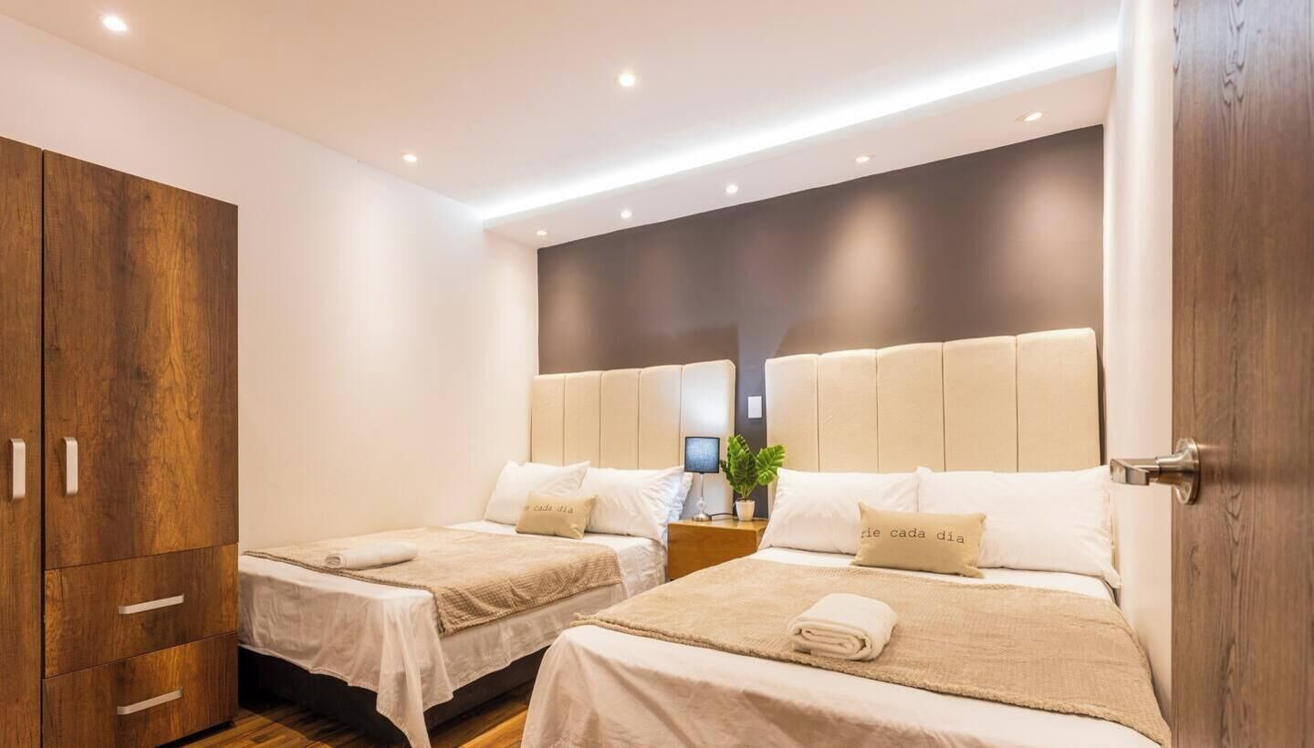 A hotel room with two beds, a wardrobe, and a bedside table with a lamp.