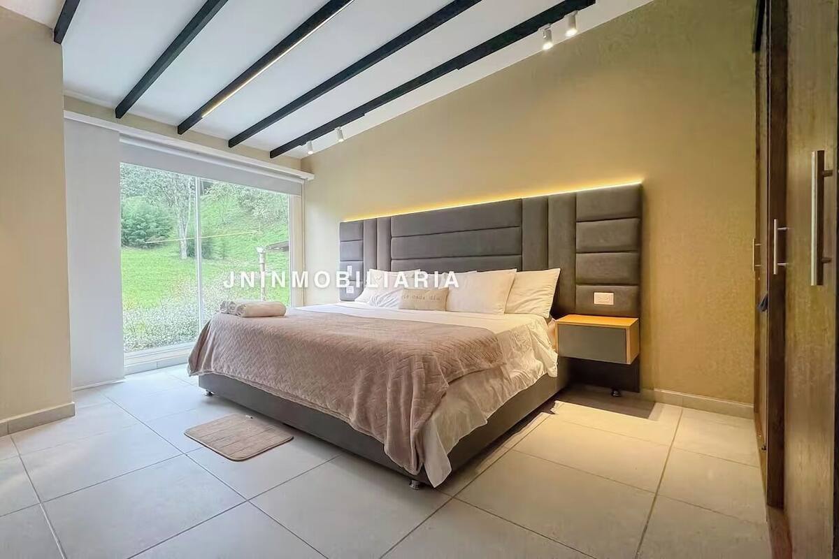 Quarto com cama grande, um banquinho, mesa de cabeceira e janela com vista para área verde.