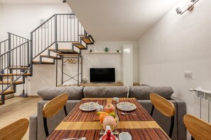 A modern living room with a dining table set for two, a sofa, a TV, and a staircase.