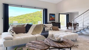 Ein modernes Wohnzimmer mit einem großen Sofa, zwei runden Couchtischen und Blick auf einen üppigen, grünen Hang durch große Fenster.